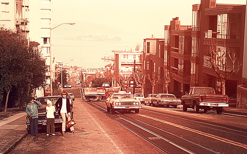 Bild i 80-tals stil med tre personer framför en bil brevid en trafikerad väg